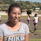 Woman in grey t-shirt speaking with young girls on the soccer field behind her.